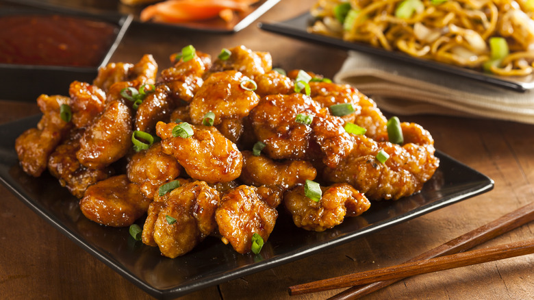 Orange chicken placed on a square black plate with chopsticks on a wooden table with Chinese noodles in the background