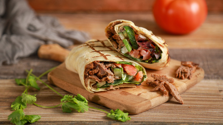 View of a shawarma wrap sliced in half and placed on a wooden board along with vegetables and garnishes